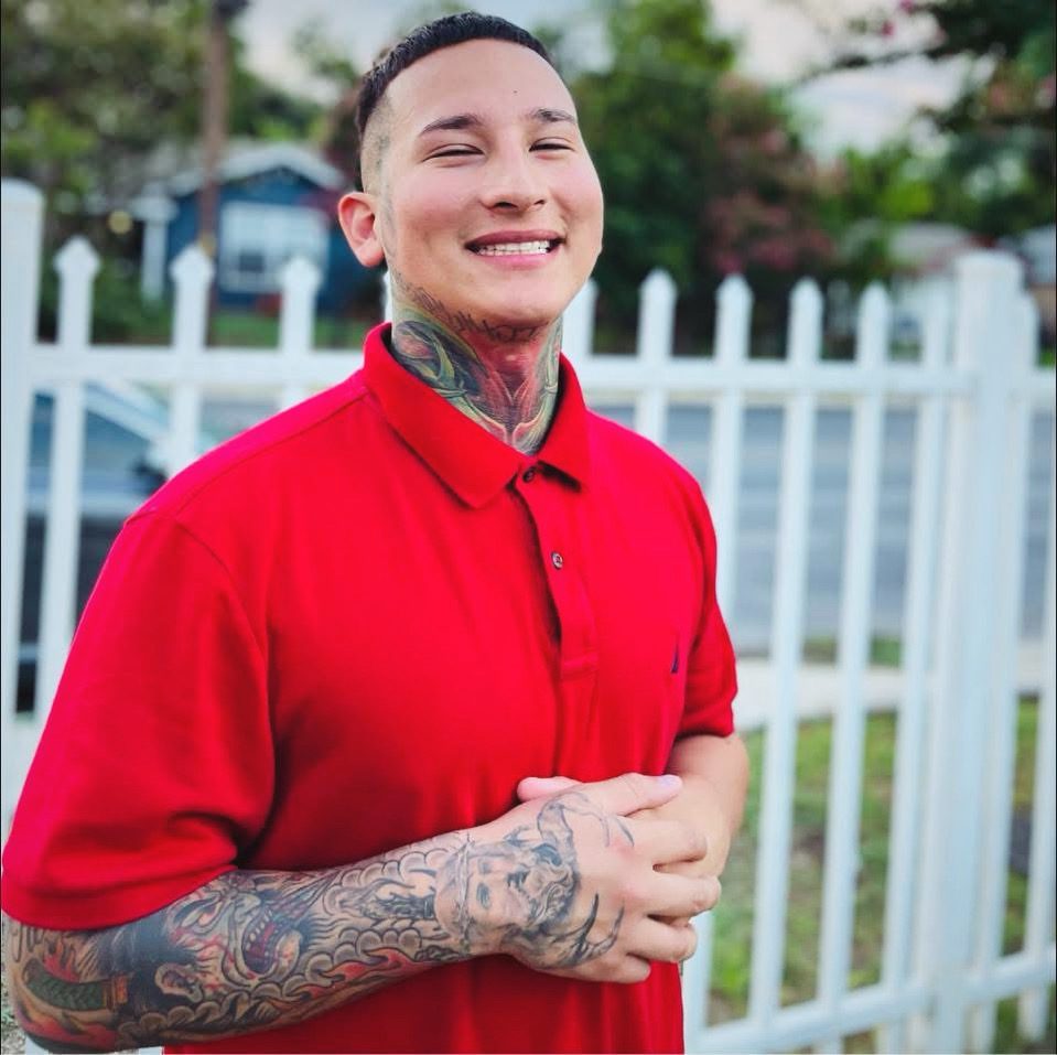 SAWEDOFF in a red polo, tattooed neck and arms, smiling in front of a white picket fence